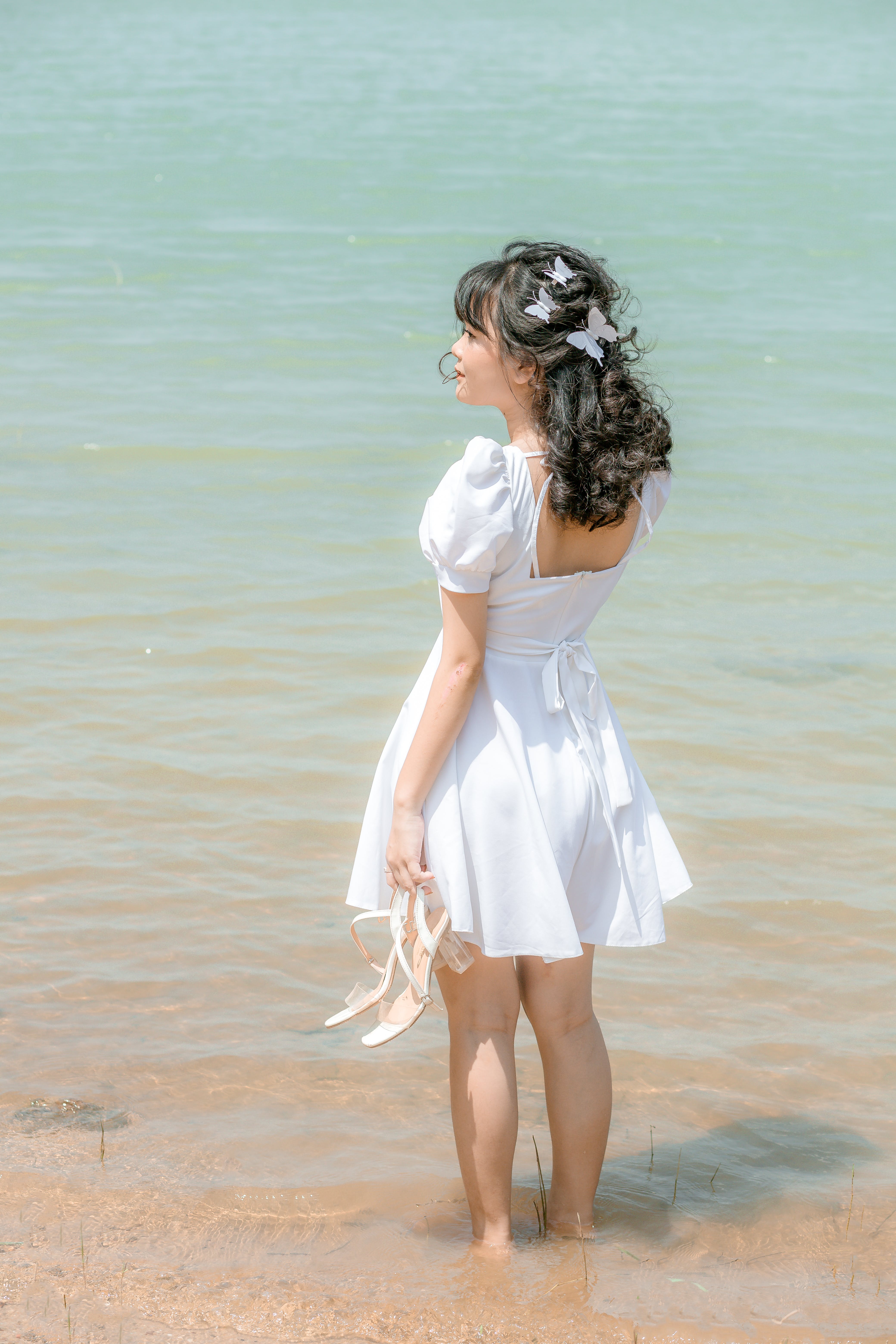 Free Back view full body of female in white elegant dress with bare back with shoes in hand standing on sand in sea Stock Photo